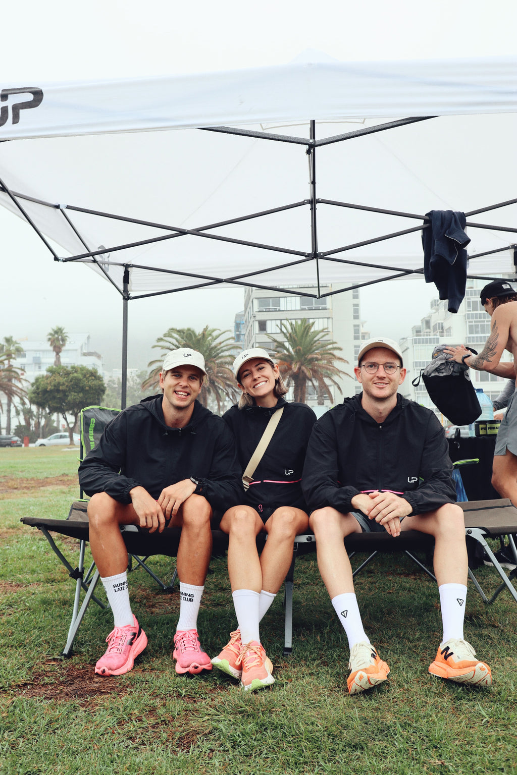 Three people sitting under a tent wearing UP Active running caps and a running windbreaker with white socks and running shoes. The caps have the text 'SHOW UP' on the back. They are doing a running event called the 4x4x48 Challenge.